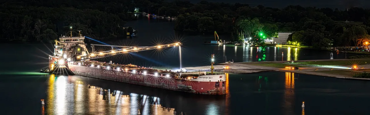 The bulk carrier Whitefish Bay lit up alongside the dock at the Port of Toronto at night, its unloading boom raised over the pier and the harbour lights reflected in the water.
