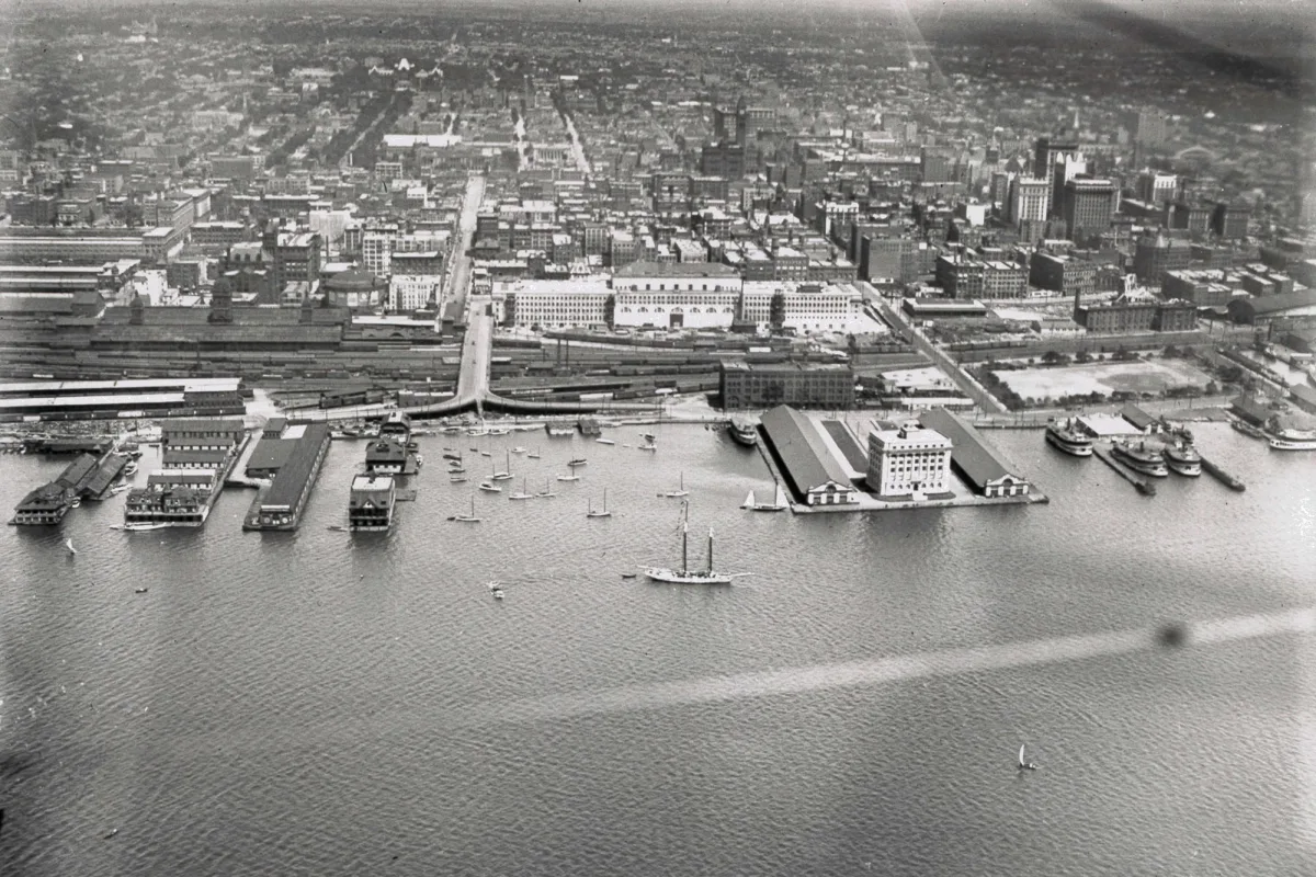 A historic black and white aerial photograph of Toronto Harbour, with ferries and a schooner on the water, the Toronto Harbour Commission building on its pier, and the city beyond.