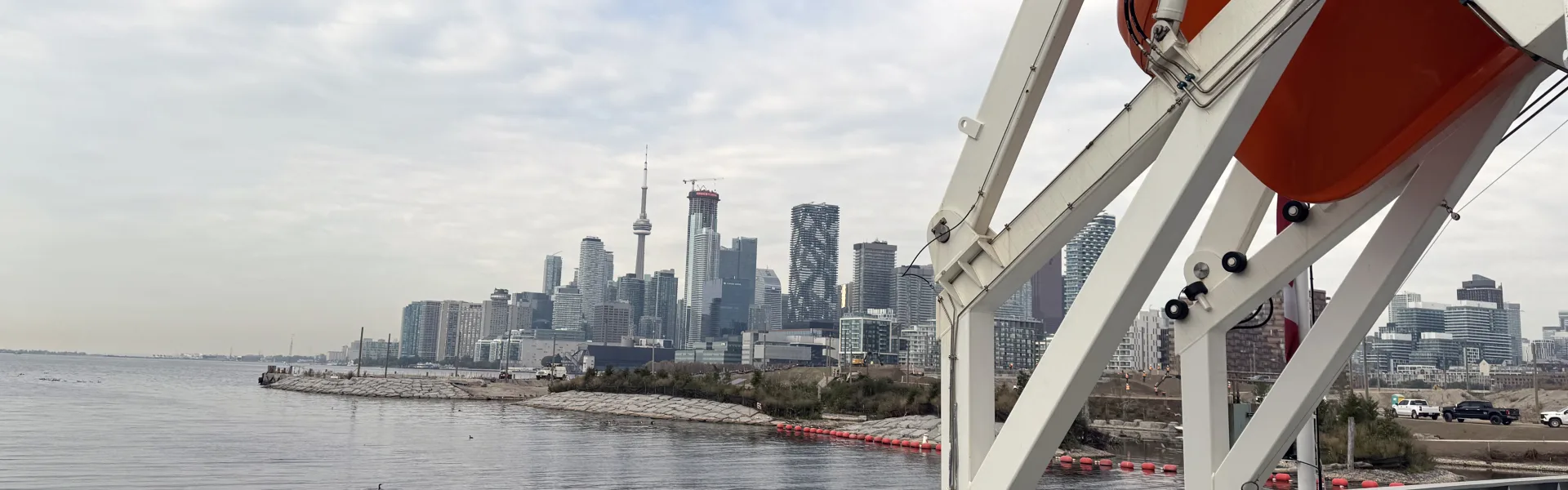 The deck of a Toronto Port Authority vessel, with mooring bollards and a lifeboat in the foreground and the Toronto skyline across the harbour.
